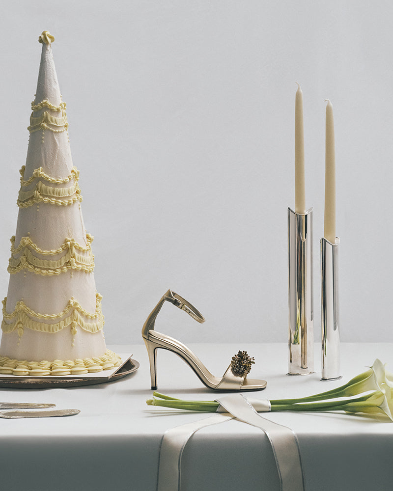 Decorative cake, gold high heel shoe, silver candle holder, and white flowers on a light gray surface.